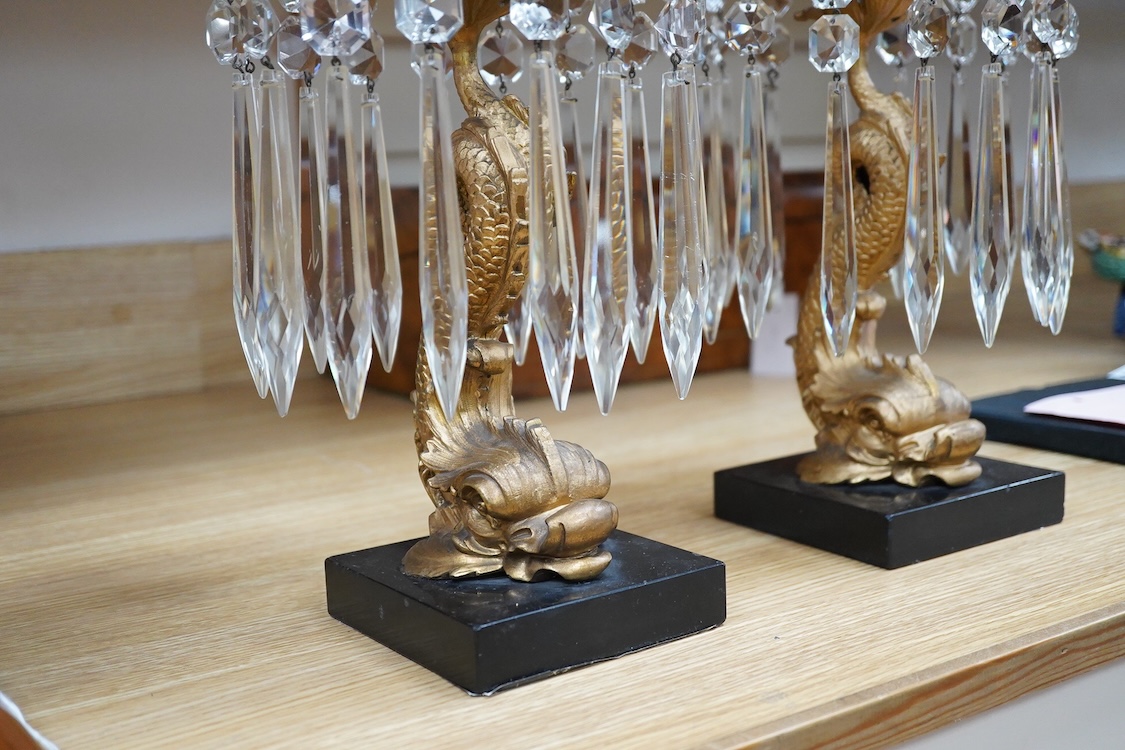 A pair of William IV gilt metal candlesticks, the stems formed as dolphins, with glass lustre drops, raised on square slate bases, 37cm high